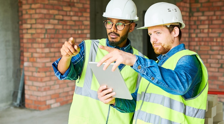 Two men in safety vests and hard hats pointing at something while holding a tablet device