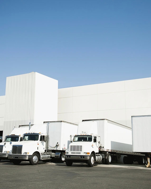 Three white package delivery trucks parked outside a white warehouse building by the loading bay.