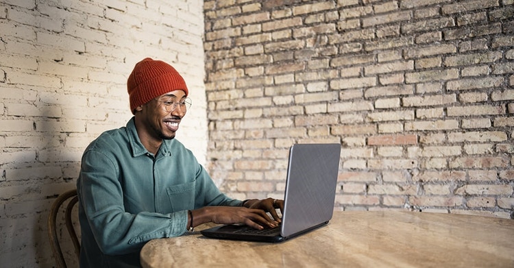 Man in a red beanie and green shirt sitting at a table using his laptop with brick wall background