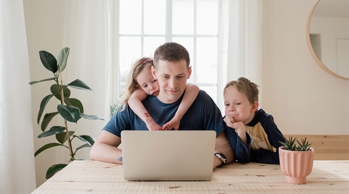A parent reviewing a house rental application with their laptop at a table while two of their children watch