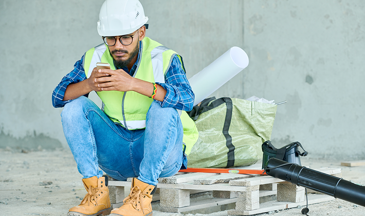 Male construction worker sitting on a wood pallet while looking at his phone