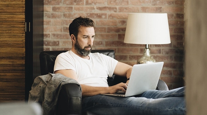 Man with a beard sitting comfortably in an airmchair couch using his laptop computer